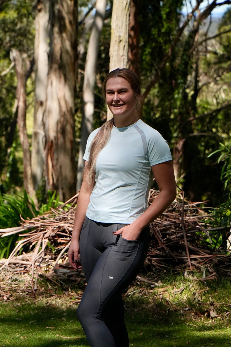 Model wearing Wilma Sport Technical Fit Tee in Sky Blue and Classic Hybrid Breeches in Slate Grey in a wooded area.